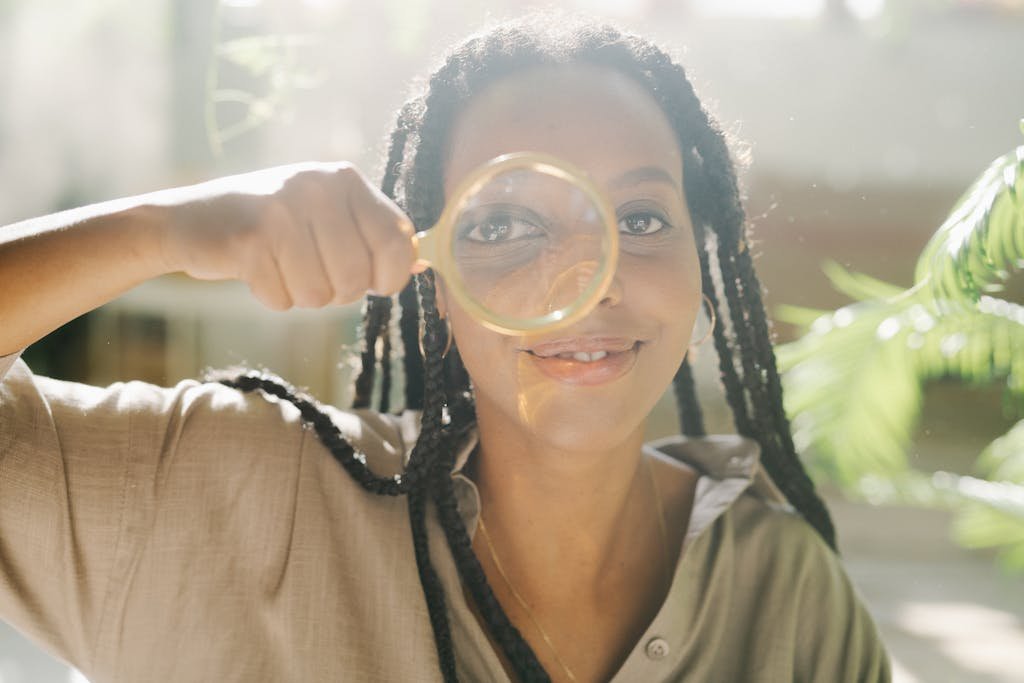 A cheerful woman with braided hair smiles as she holds a magnifying glass indoors.