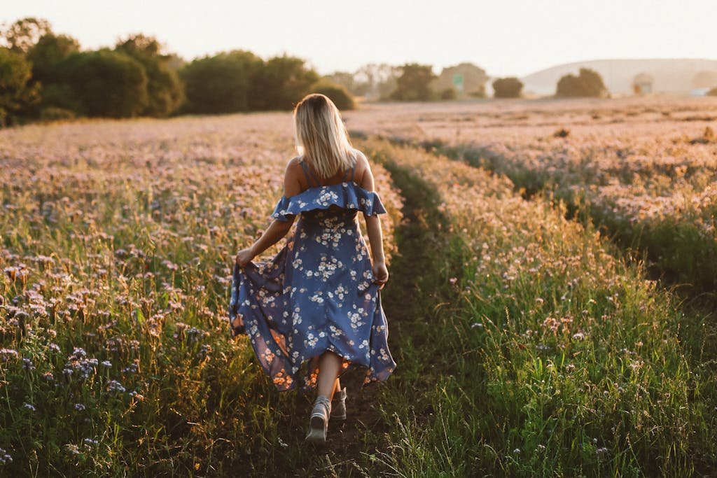 A woman walking in a floral dress through a bright flower field during sunset, showcasing tranquility and nature.