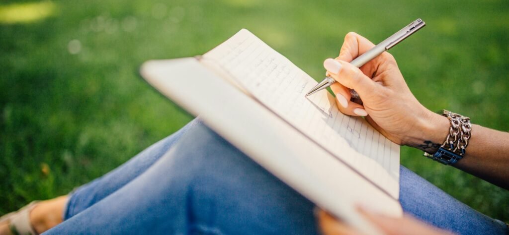 Person writes in a notebook on a sunny day. Focus on hands and pen outdoors.