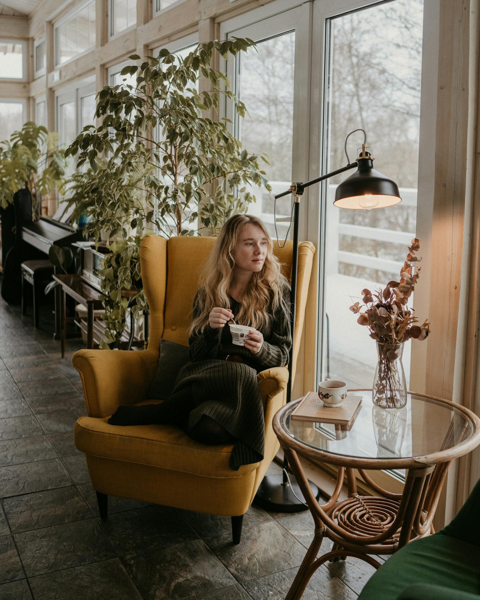 Woman sitting in a cozy armchair by a window, surrounded by houseplants, enjoying a quiet moment indoors.