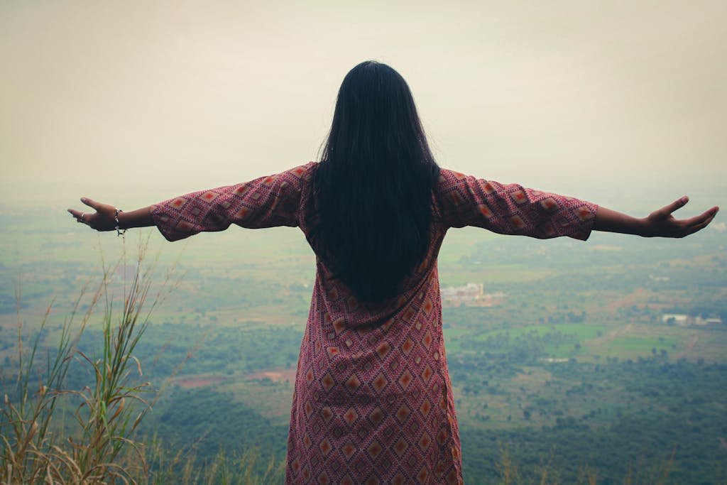Back view of woman in pink dress standing on mountain, arms spread wide, embracing nature.